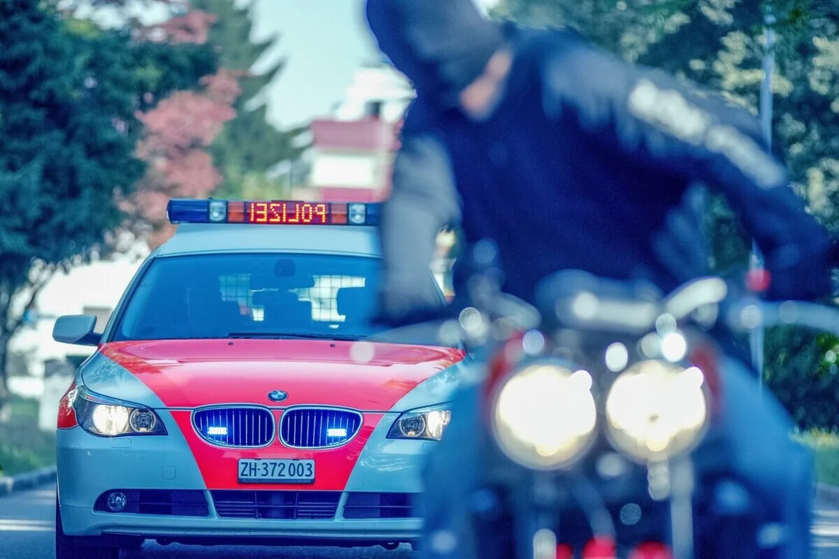 Ein Polizeiwagen mit roten und weißen Farben und eingeschalteten Lichtern fährt auf einer Straße. Ein unscharfer Motorradfahrer ist im Vordergrund sichtbar.