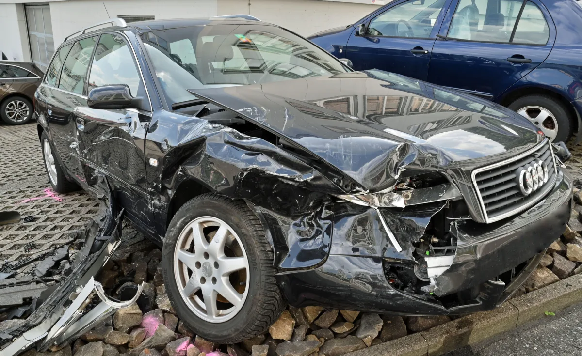 Ein schwarzer Audi mit schweren Schäden an der Vorderseite steht auf einem Parkplatz. Der Vorderkotflügel ist eingedrückt und der Stoßfänger hängt herab.