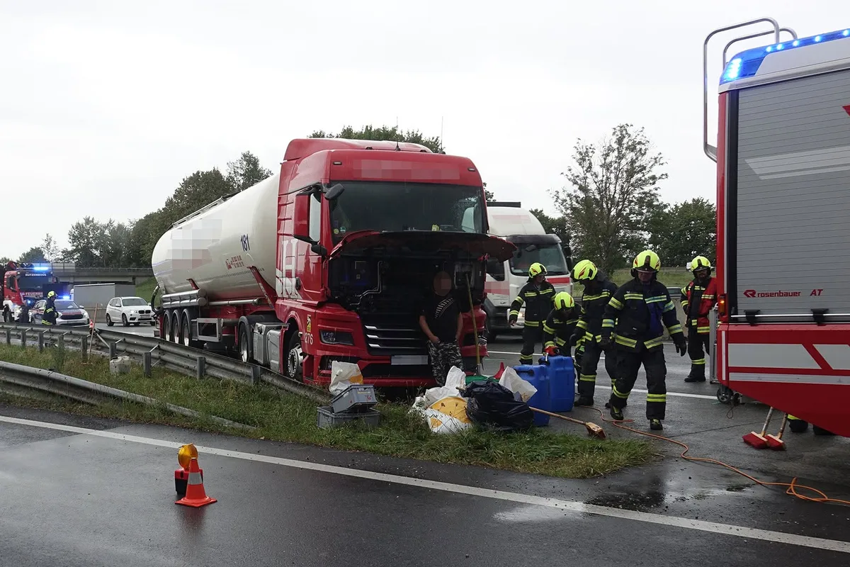 Ein roter Lastwagen steht am Straßenrand mit geöffnetem Motorraum. Mehrere Feuerwehrleute in gelben Helmen arbeiten daneben.