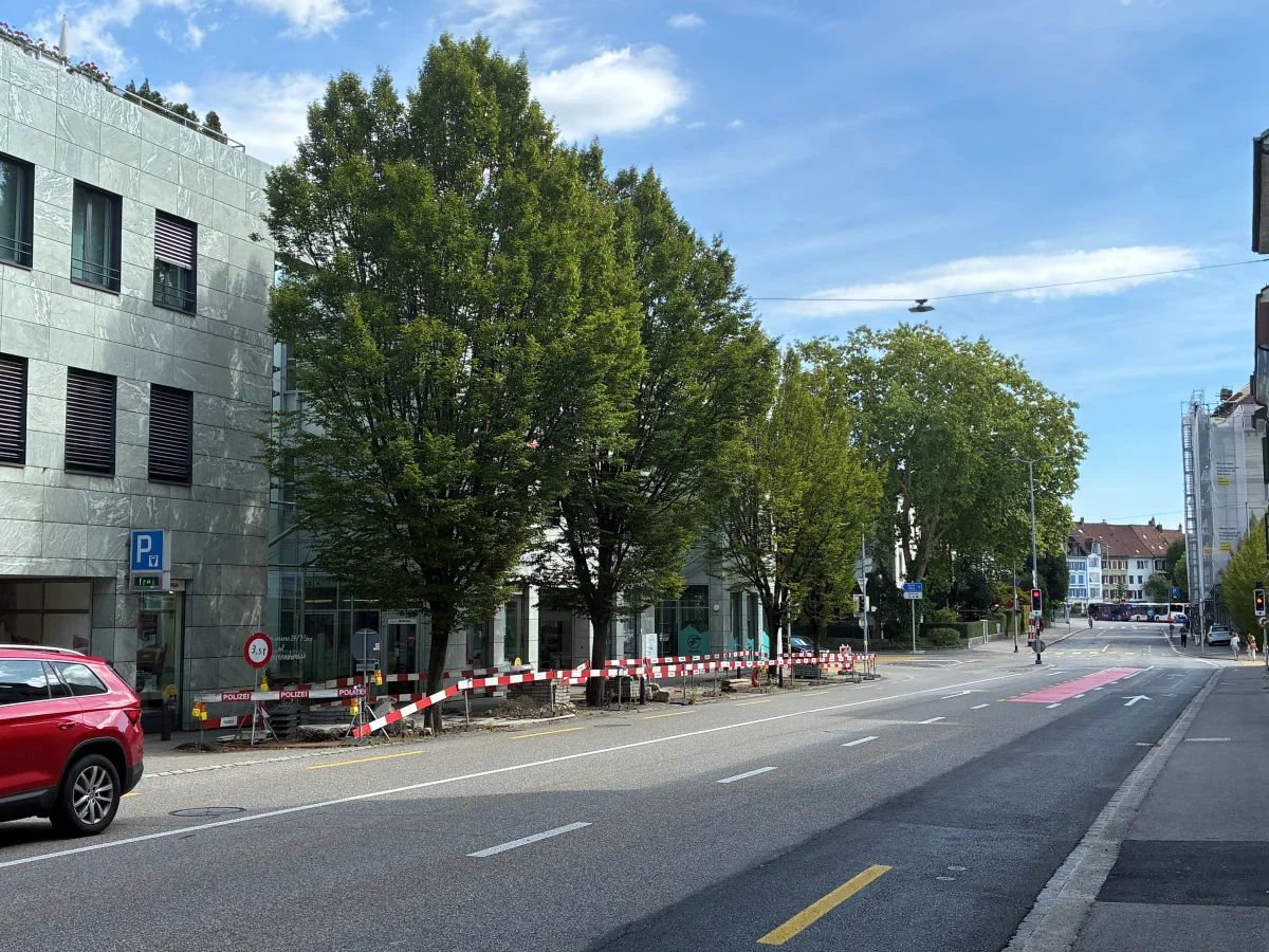 Ein rotes SUV steht vor einer Baustelle, die mit Polizeiabsperrungen gesichert ist. Der Himmel ist bewölkt, Gebäude und Bäume säumen die Straße.