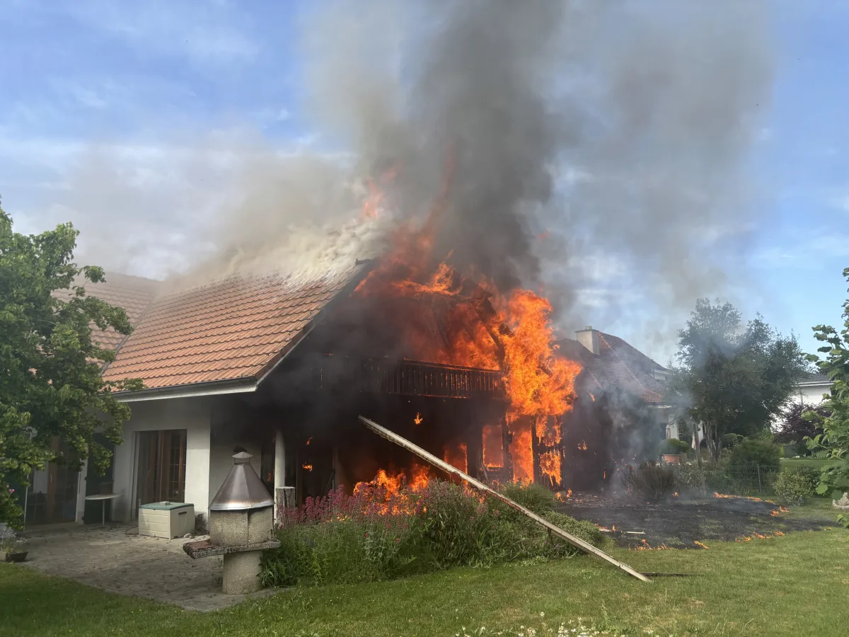 Ein Wohnhaus ist stark in Brand geraten, Flammen schlagen aus dem Dach und dichter Rauch steigt auf, während ein Garten im Vordergrund zu sehen ist.