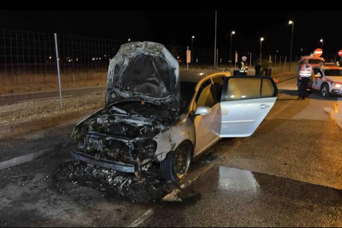 Ein silbernes Auto mit offenem, ausgebranntem Motorraum steht in einer Nachtaufnahme am Straßenrand. Im Hintergrund sind Polizisten und ein Einsatzfahrzeug sichtbar.