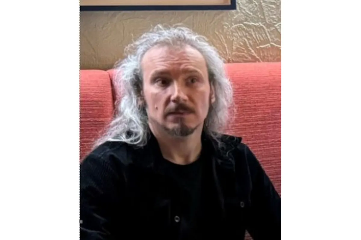 Ein Mann mit langen grauen Haaren und Bart sitzt in einem Sessel mit rotem Stoffbezug. Im Hintergrund eine beige Wand.