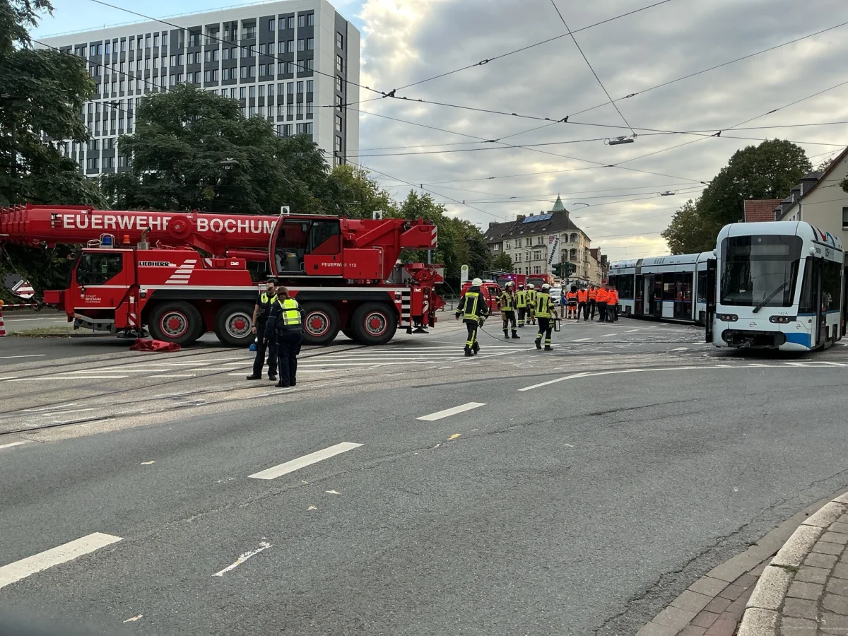 Ein großes rotes Feuerwehrfahrzeug von 'Feuerwehr Bochum' steht auf der Straße. Einsatzkräfte sind um eine entgleiste Straßenbahn herum versammelt.