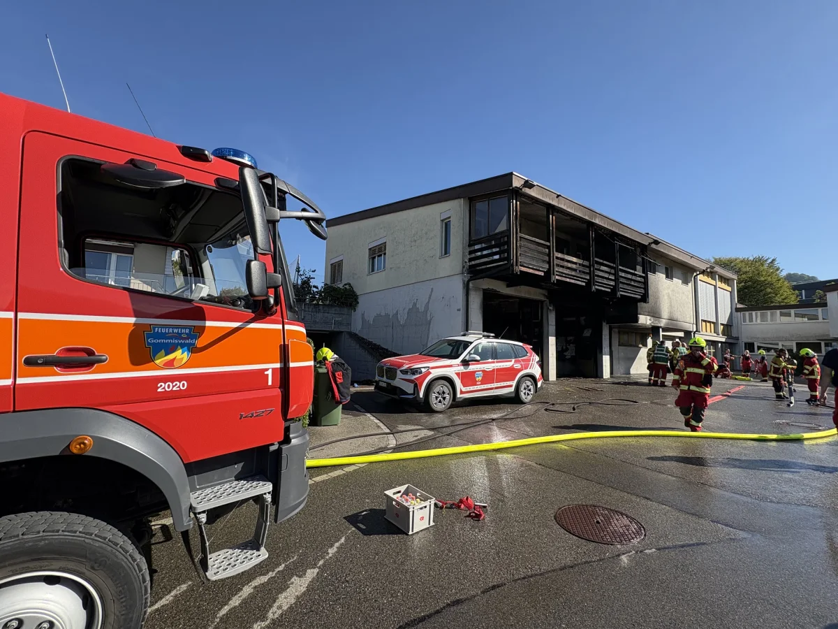 Ein rotes Feuerwehrfahrzeug mit der Aufschrift 'Feuerwehr Gommiswald' steht vor einem angebrannten Gebäude. Mehrere Feuerwehrleute in Schutzkleidung arbeiten vor Ort.