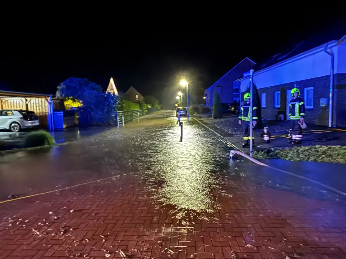 Zwei Feuerwehrleute stehen in einer nachts überfluteten Wohnstraße. Das Wasser bedeckt die Pflastersteine, während die Straßenlaternen beleuchtet sind.