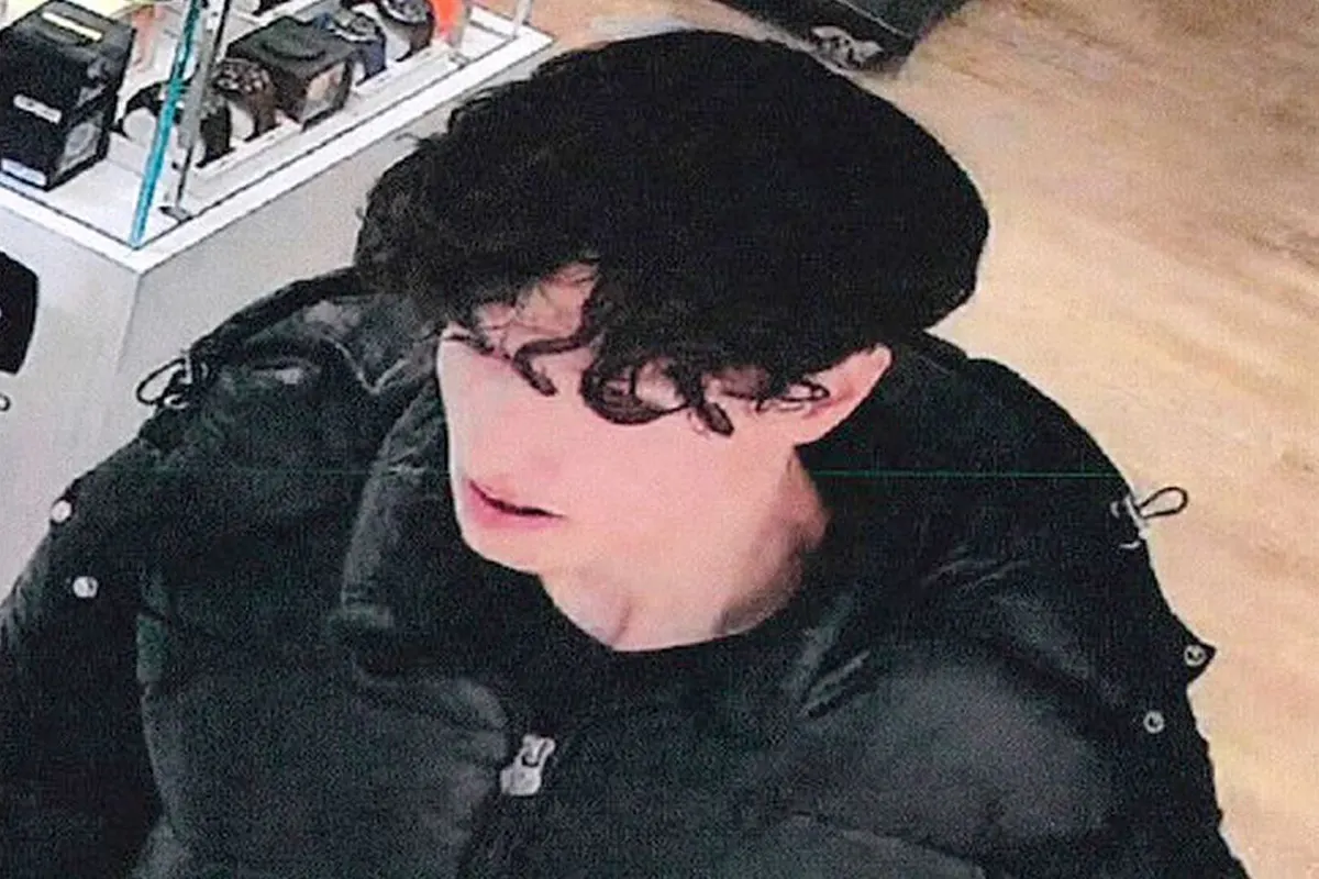 Eine unbekannte junge Person mit lockigem Haar in einer dunklen Jacke steht in einem Laden. Die Umgebung ist ein Verkaufsraum mit Regalen.