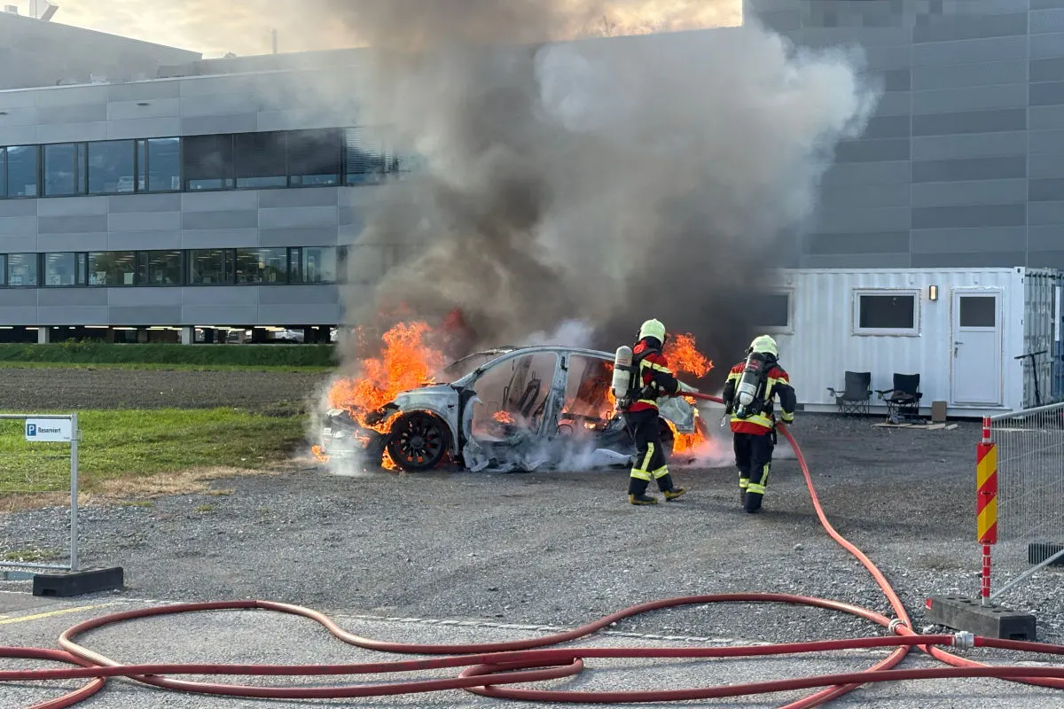 Zwei Feuerwehrleute mit Atemschutzgeräten löschen ein brennendes Auto auf einem Parkplatz vor einem Bürogebäude.