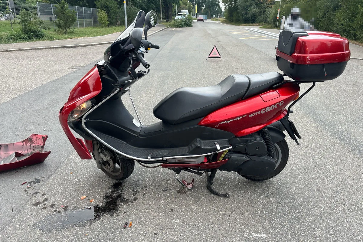 Ein roter Motorroller mit sichtbaren Frontschäden steht auf einer asphaltierten Straße. Im Hintergrund sind eine Warntafel und Vegetation zu sehen.