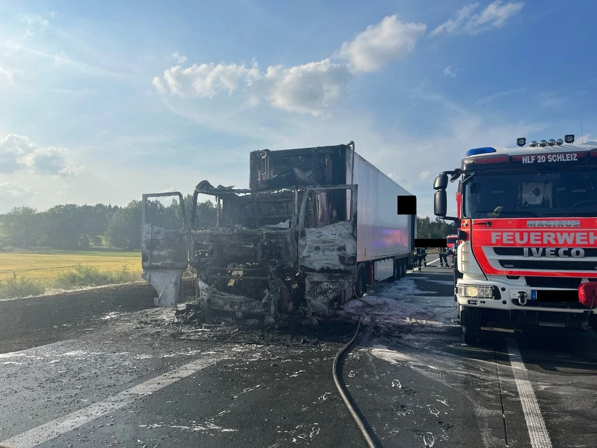 Ein ausgebrannter Lastwagen steht auf einer Autobahn, während ein Feuerwehrfahrzeug daneben parkt. Der Himmel ist leicht bewölkt.