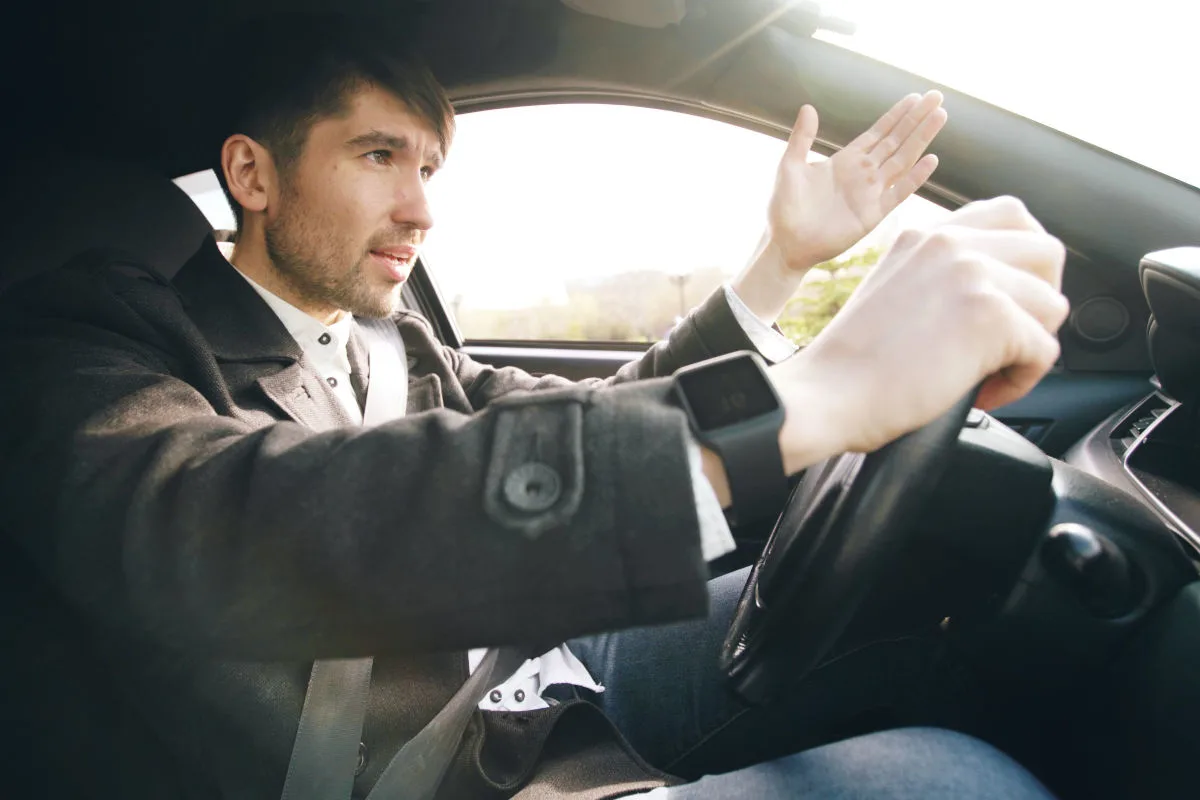 Ein junger Mann im Auto gestikuliert mit der linken Hand, während er fährt. Er trägt einen Mantel und eine Smartwatch.