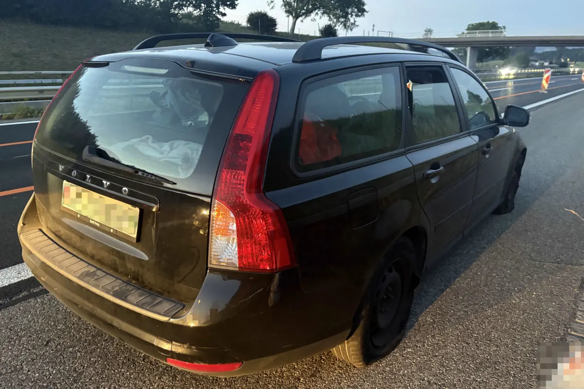 Ein schwarzer Volvo Kombi steht am Straßenrand einer Autobahn. Im Hintergrund sind Bäume und eine Brücke erkennbar.