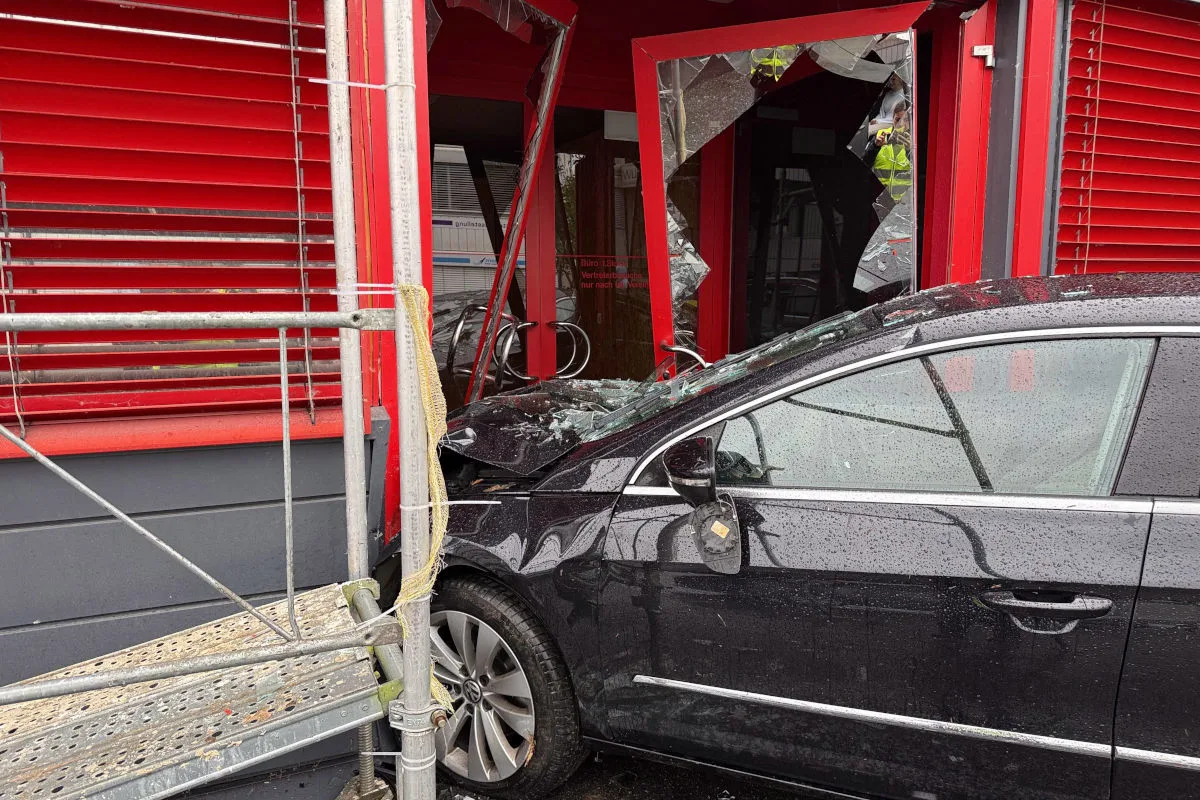 Ein schwarzes Auto ist in die Fensterfront eines roten Gebäudes gefahren, zerbrochenes Glas ist sichtbar.