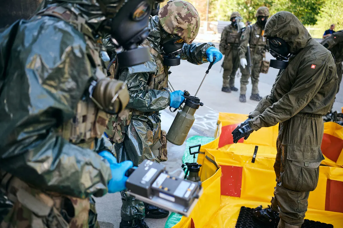 Mehrere Personen in Schutzanzügen und Gasmasken nehmen an einer Dekontaminationsübung teil. Einer sprüht eine Flüssigkeit aus einem Behälter.