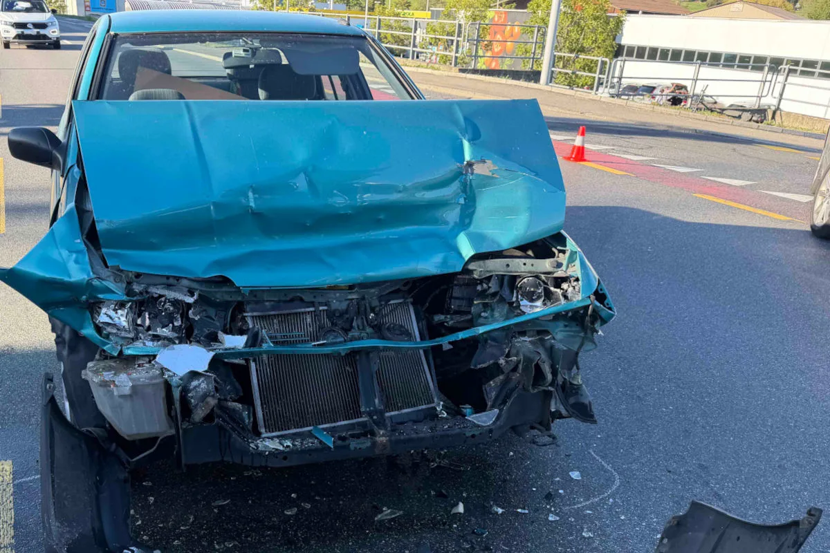 Ein grünes Auto mit schwer beschädigter Front steht auf einer Straße. Im Hintergrund sind Verkehrszeichen und andere Fahrzeuge sichtbar.