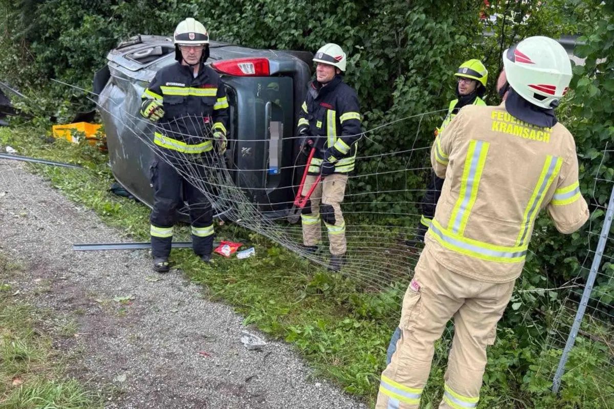 Feuerwehrleute arbeiten an einem umgestürzten Auto, das neben einem Zaun liegt. Sie tragen Schutzkleidung und Helme.