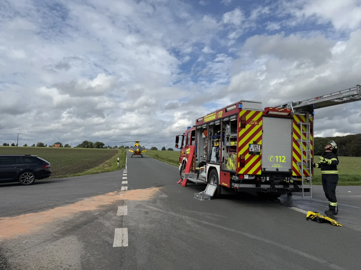Ein Feuerwehrfahrzeug mit geöffnetem Equipment steht auf einer Landstraße. Ein Rettungshubschrauber befindet sich auf der Straße.