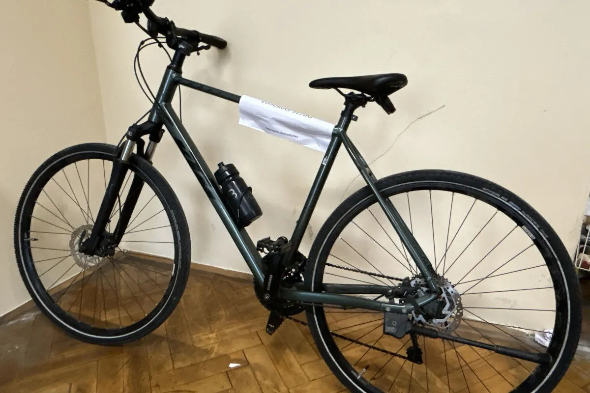 Ein grünes Fahrrad steht in einem Raum an eine weiße Wand gelehnt. Am Rahmen hängt ein weißer Zettel.