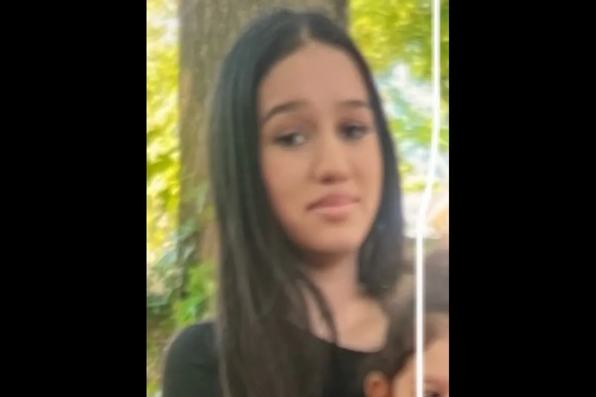 Das Bild zeigt eine junge Person mit langen Haaren im Freien vor einem Baum. Die Person blickt nach vorne.