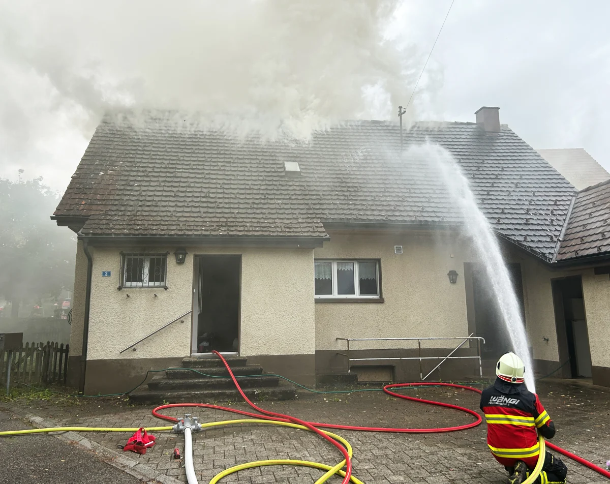 Ein Feuerwehrmann in Schutzkleidung sprüht Wasser auf ein Wohnhausdach, aus dem Rauch aufsteigt.