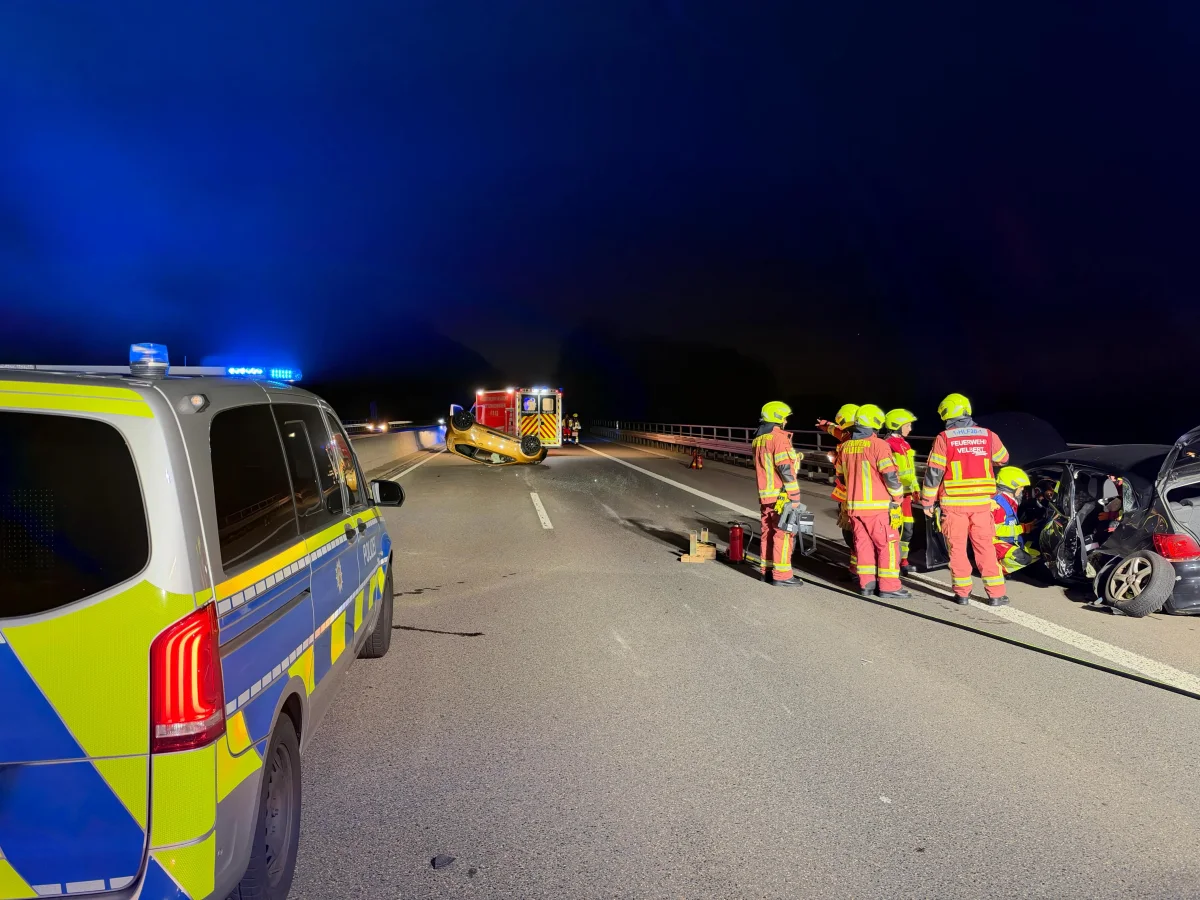 Eine Gruppe von Feuerwehrleuten steht neben einem beschädigten Fahrzeug auf einer Autobahn bei Nacht. Ein Polizeifahrzeug und ein weiteres Einsatzfahrzeug sind ebenfalls anwesend.