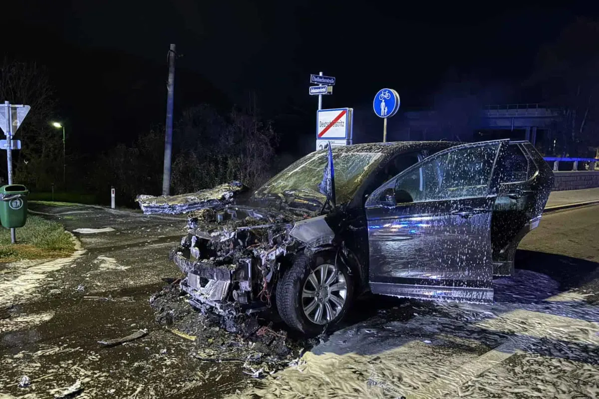 Ein schwarzes Auto mit geöffneter Fahrertür und stark beschädigter Front auf einer nächtlichen Straße mit Schildern im Hintergrund.