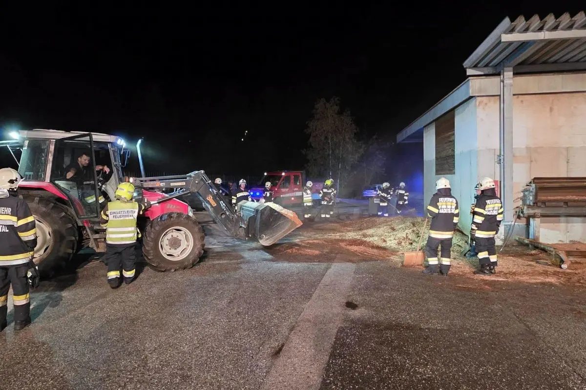 Mehrere Feuerwehrkräfte stehen nachts um einen Traktor, der nahe eines Gebäudes geparkt ist. Sie sind mit Aufräumarbeiten beschäftigt.