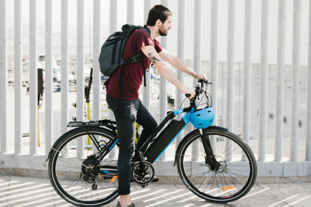 Ein Mann mit rotem T-Shirt und Rucksack steht neben einem blauen Fahrradhelm und einem Fahrrad vor einem Metallzaun.