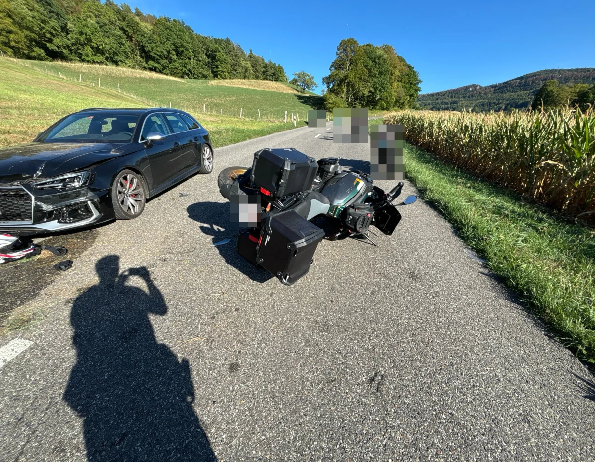 Ein gestürztes Motorrad liegt auf der Straße neben einem schwarzen Auto. Im Hintergrund sind Bäume und Felder sichtbar.