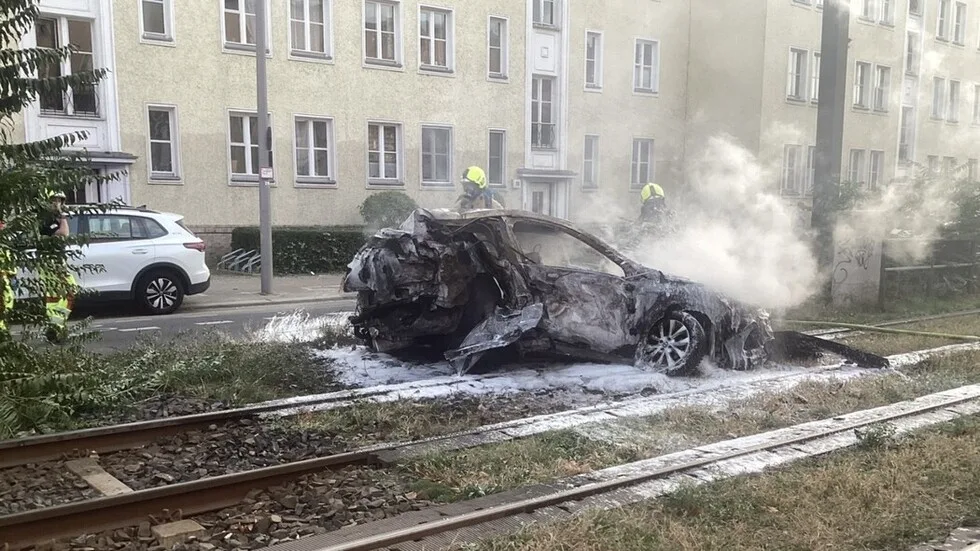 Ein ausgebranntes Auto liegt auf Bahngleisen, umgeben von Feuerwehrleuten, die den Brand löschen. Im Hintergrund ist ein Wohngebäude zu sehen.