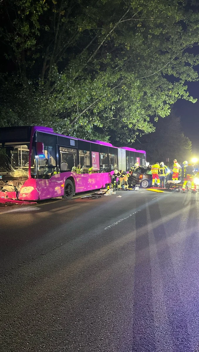 Ein Bus und ein Pkw stehen nach einem Unfall auf einer Landstraße, umgeben von Feuerwehr- und Rettungskräften bei Nacht.