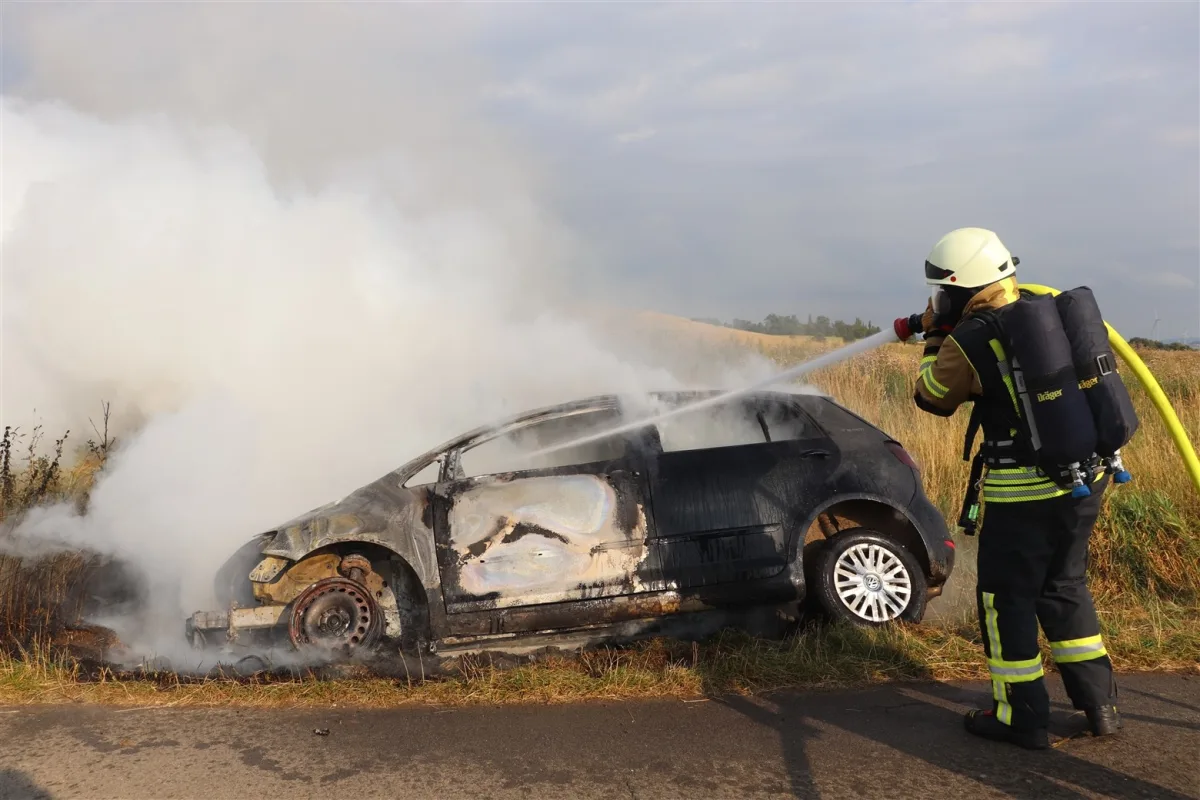 Ein Feuerwehrmann in voller Ausrüstung löscht mit einem Wasserschlauch einen brennenden PKW, der stark vernebelt ist.
