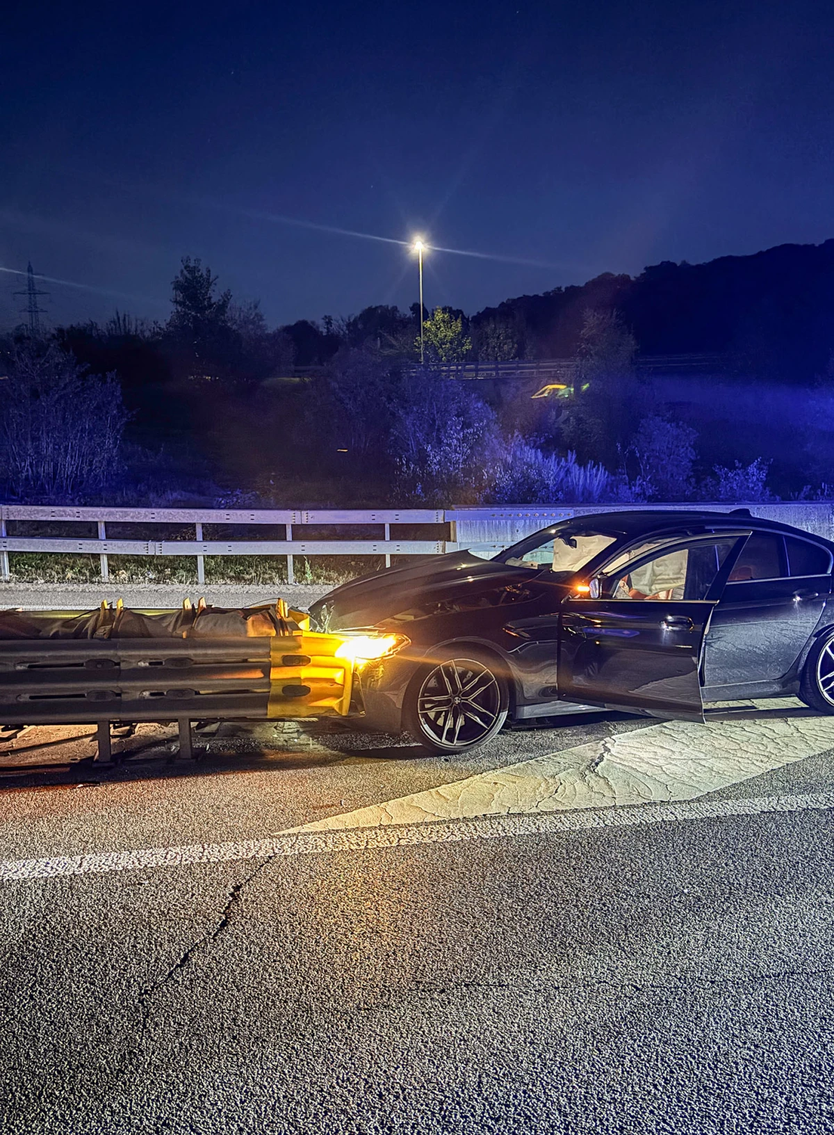 Ein dunkles Auto steht nachts auf einer Straße mit der Front in einer Leitplanke. Die Szene ist beleuchtet.