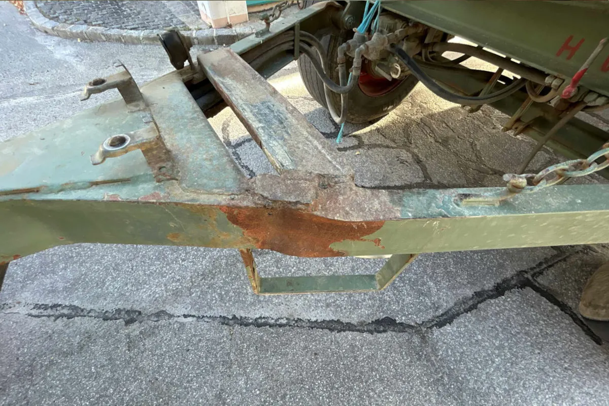 Ein verrosteter Anhängerrahmen auf einer asphaltierten Straße. Der Fokus liegt auf den sichtbaren Schäden.