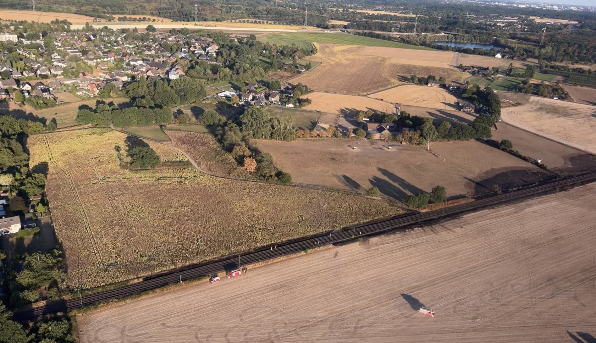 Eine ländliche Landschaft aus der Vogelperspektive. Einsatzfahrzeuge stehen neben Bahngleisen entlang eines großen Feldes.