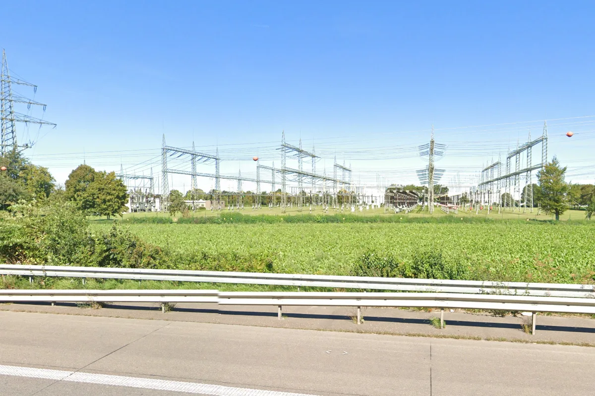 Eine Umspannstation ist hinter einer grünen Wiese und einer Straße sichtbar. Der Himmel ist klar und blau, was auf gutes Wetter schließen lässt.