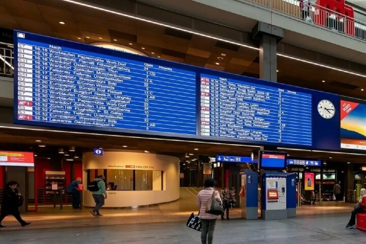Ein großer digitaler Anzeigebildschirm im Bahnhof zeigt verschiedene Zugzeiten und -ziele. Menschen stehen oder gehen in der Halle darunter.
