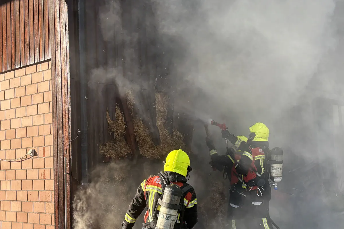 Zwei Feuerwehrleute in gelben Helmen löschen mit einem Schlauch ein Feuer in einer stark verrauchten Scheune. Dichtes Stroh ist sichtbar.