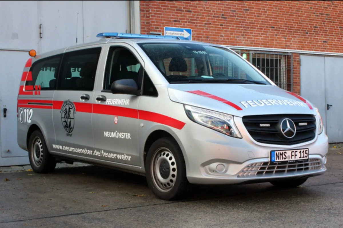 Ein silberner Mercedes-Van der Feuerwehr Neumünster steht geparkt vor einem Backsteingebäude. Auf der Seite sind rote Streifen und das Stadtwappen zu sehen.