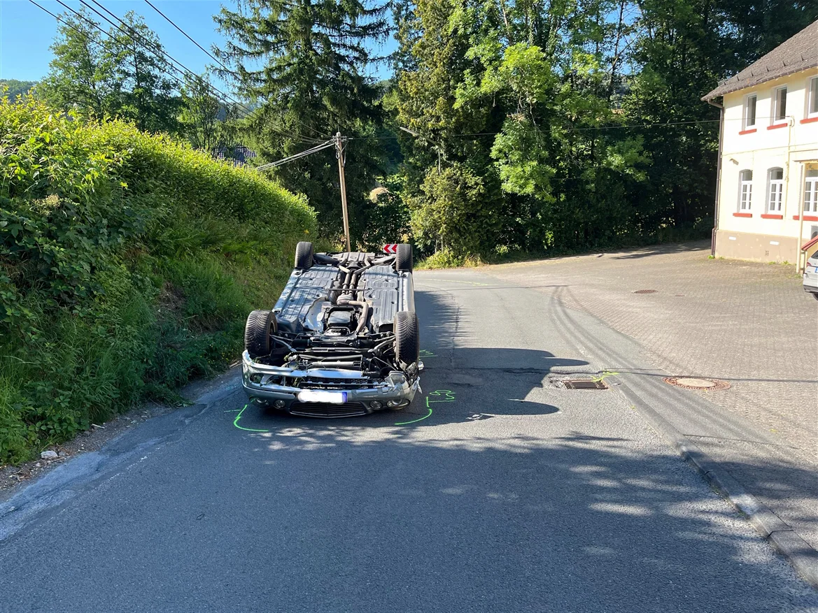 Ein Auto liegt umgestürzt auf dem Dach auf einer Straße in einem Wohngebiet. Im Hintergrund sind Bäume und ein Wohnhaus zu sehen.