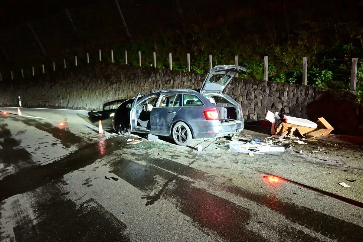 Ein graues Auto steht beschädigt auf einer nassen Straße. Die Heckklappe ist geöffnet, und Trümmer sind auf der Fahrbahn verstreut.
