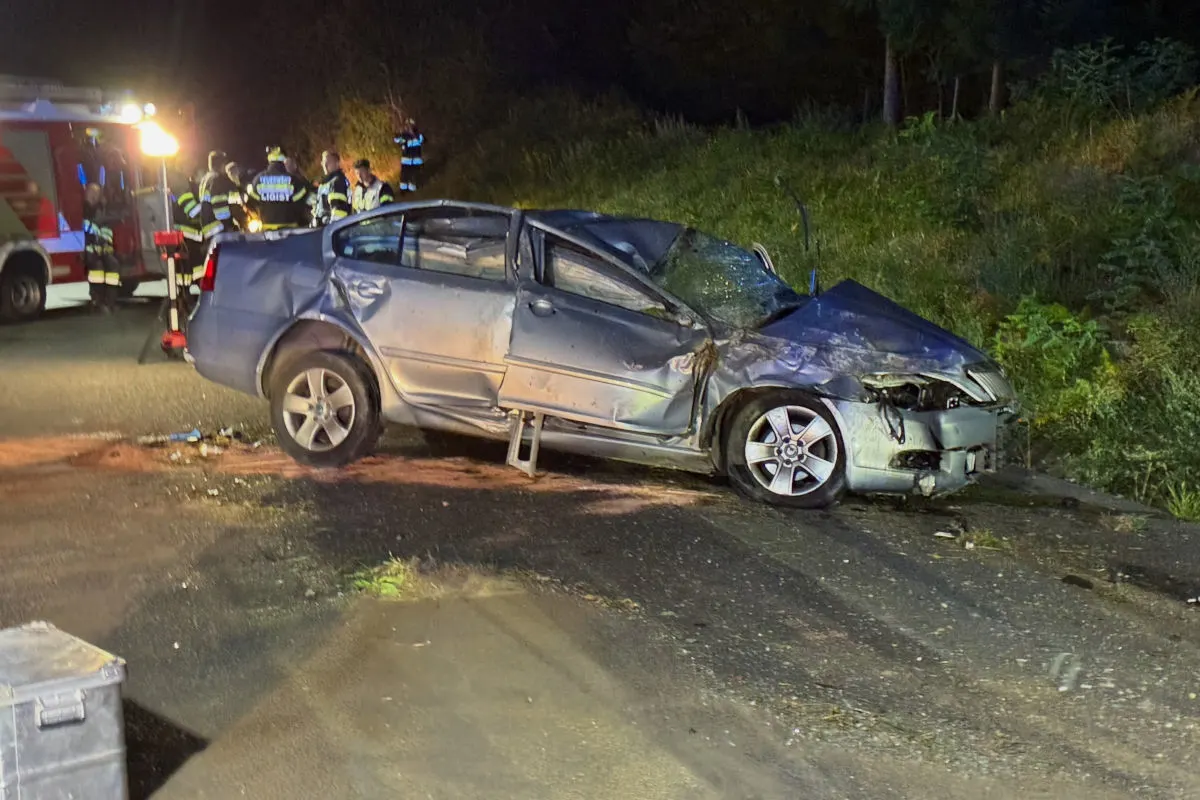 Ein silbernes Auto mit schweren Frontschäden steht am Straßenrand, umgeben von Einsatzfahrzeugen und Einsatzkräften bei Nacht.