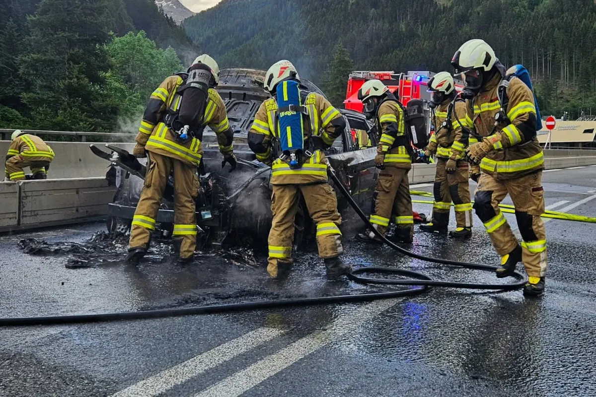Mehrere Feuerwehrleute in voller Ausrüstung löschen die Überreste eines ausgebrannten Autos auf einer Autobahn. Im Hintergrund sind Berge und Bäume zu sehen.