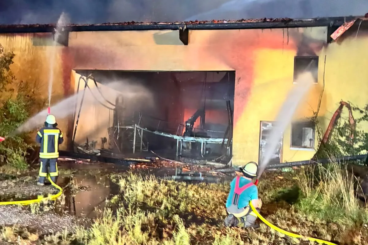 Zwei Feuerwehrleute bekämpfen nachts mit Löschschläuchen einen Großbrand in einem Gebäude. Flammen sind im Inneren zu sehen.