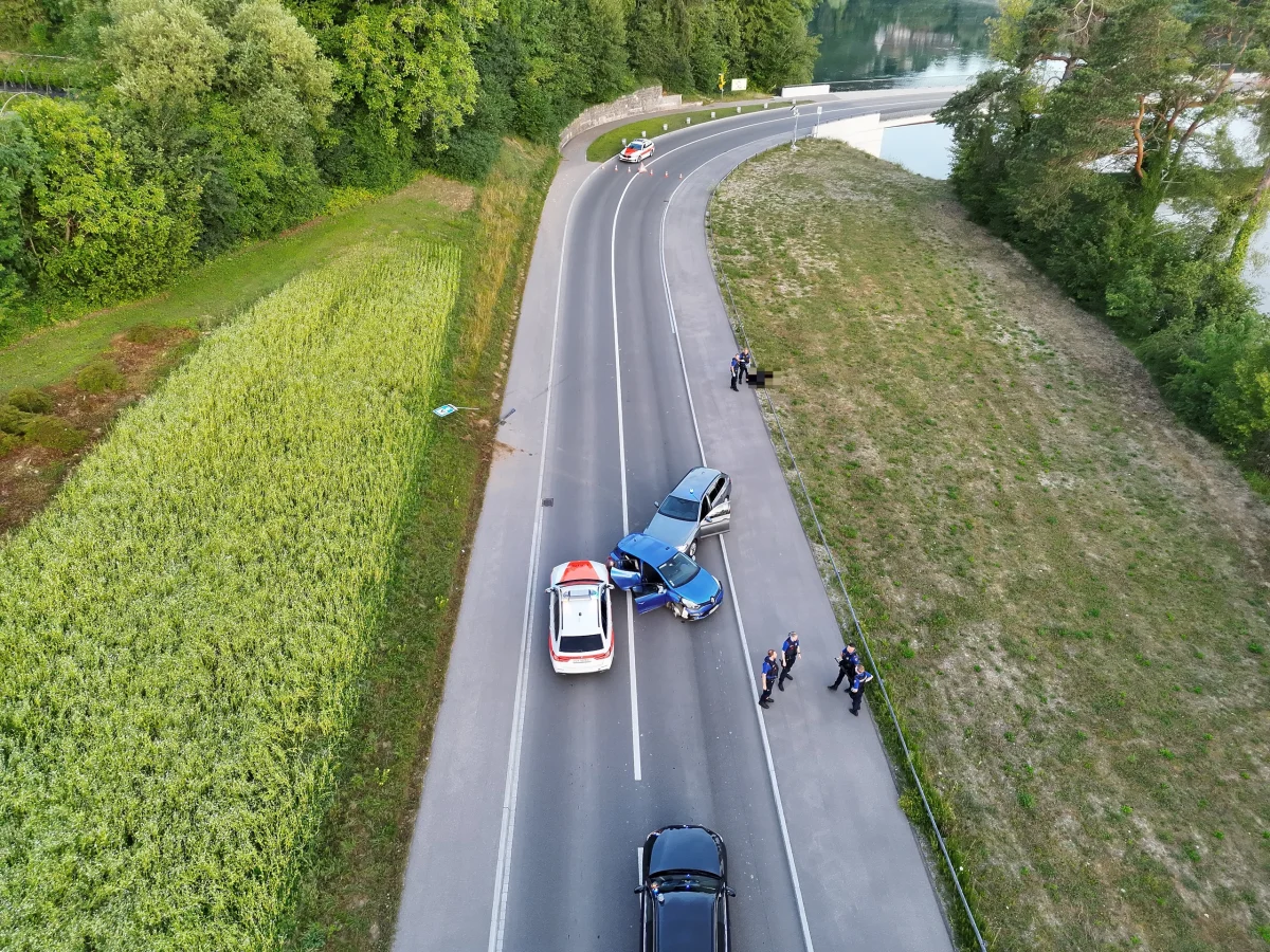 Luftaufnahme einer Landstraße mit einem blauen und einem weißen Auto, umgeben von Polizeibeamten. Das Wetter ist klar.
