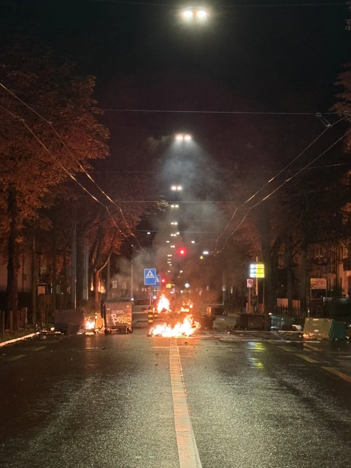Brennende Gegenstände auf einer dunklen Straße, umgeben von Bäumen, mit mehreren erleuchteten Straßenlaternen in der Mitte.