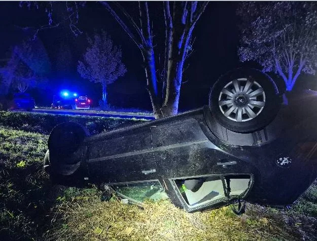 Ein Auto liegt auf der Seite im Gras. Im Hintergrund sind Polizeifahrzeuge mit eingeschaltetem Blaulicht zu sehen.