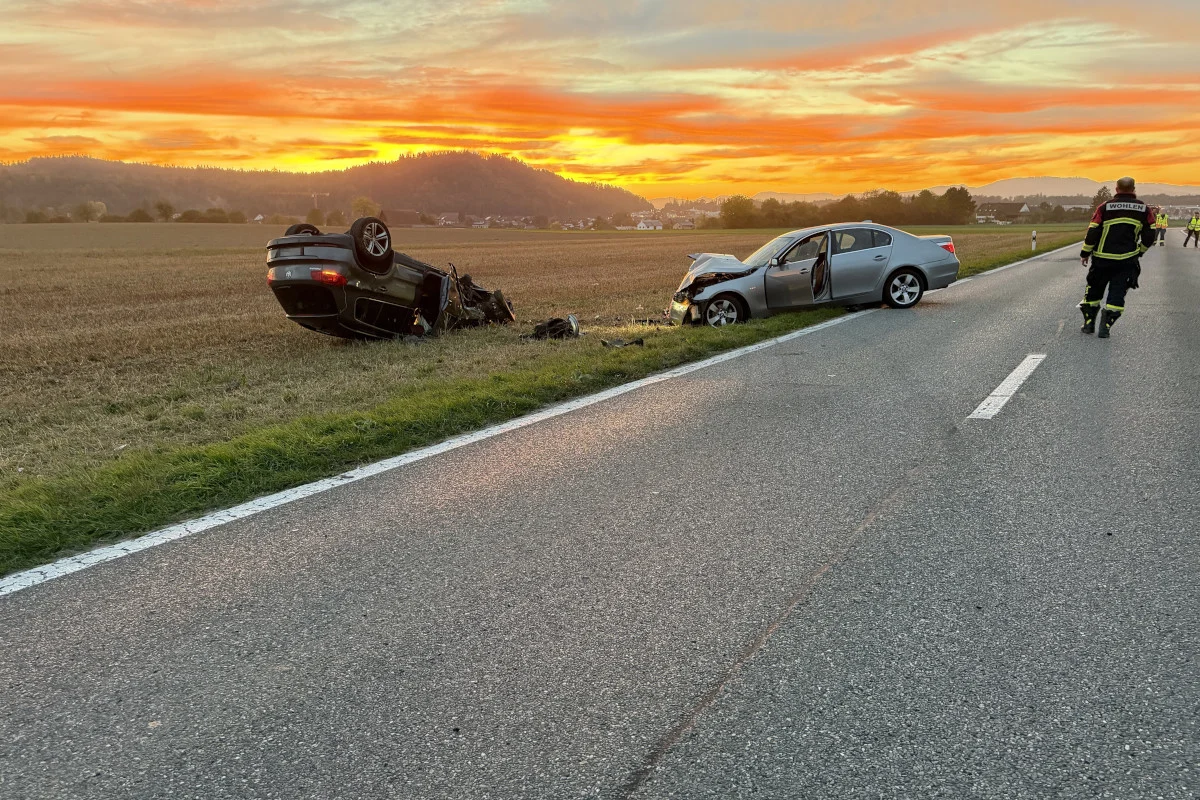 Ein Fahrzeug liegt auf dem Dach im Feld, während ein zweites beschädigtes Auto am Straßenrand steht. Am Himmel ist der Sonnenuntergang zu sehen.