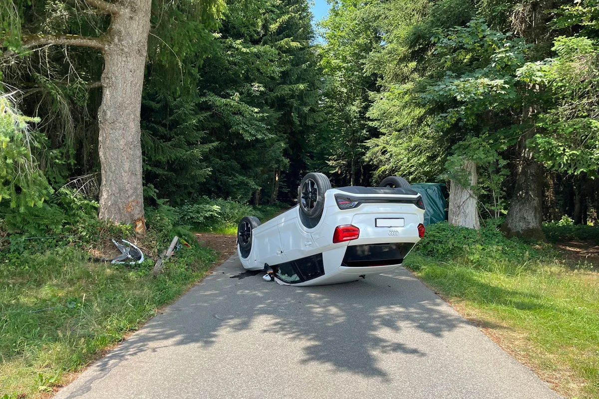Ein weißes Auto liegt auf dem Dach auf einer schmalen Straße, umgeben von Bäumen in einem Waldgebiet. Die Szene wirkt ruhig, da keine Menschen sichtbar sind.
