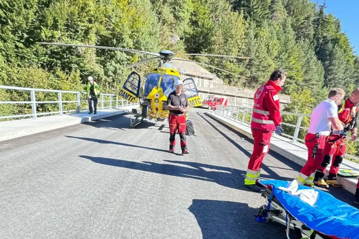 Ein gelber Rettungshubschrauber steht auf einer Straße. Um ihn herum befinden sich mehrere Rettungskräfte in roten Uniformen. Die Straße ist von grünen Wäldern umgeben.